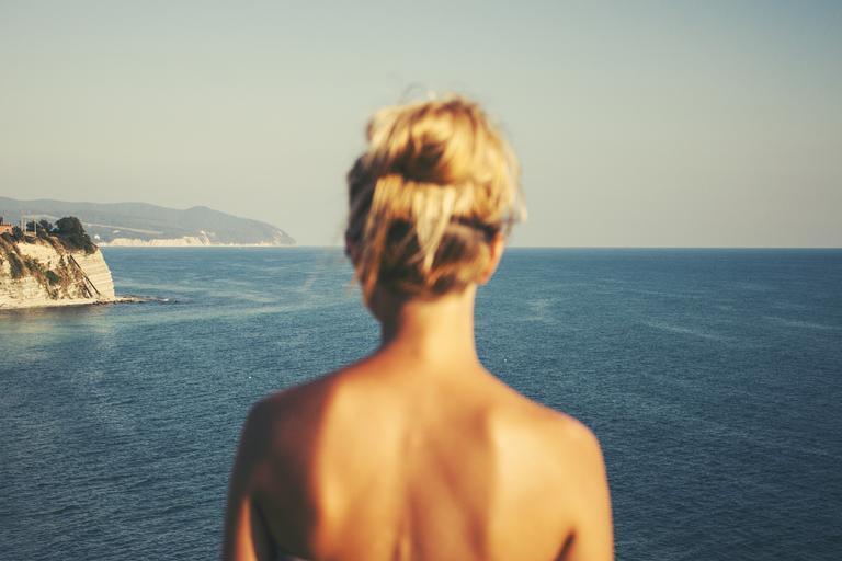 žena s blond vlasy do drdolu stojí zády a dívá se na moře, má holá záda